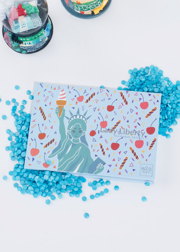 statue of liberty inspired eyeshadow palette sitting among blue sprinkles and two snow globes