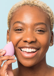 smiling girl holding a purple makeup blending sponge next to her face
